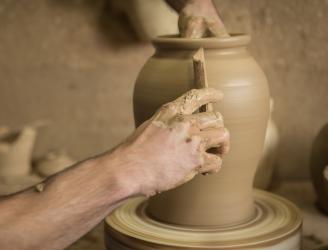 Imagen de una pieza de arcilla que va tomando forma, junto a las manos expertas de un alfarero, que la modela al torno con la ayuda de una caña para alisar la superficie.
