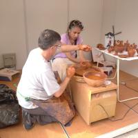 Aprendiza en el torno de alfarero con el artesano dirigiéndole como tiene que mover la pella de arcilla.